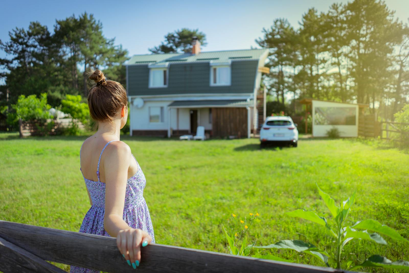 A woman in a sundress stands by a fence admiring a countryside house on a sunny day.