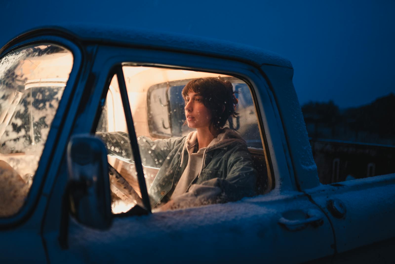 A woman sits in an illuminated, snow-covered pickup truck during a cold winter night.