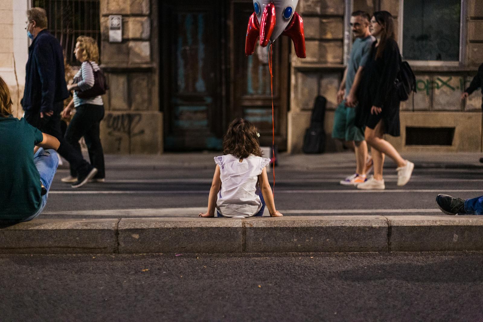 A child sits on a city street with a rocket balloon, observing passersby.