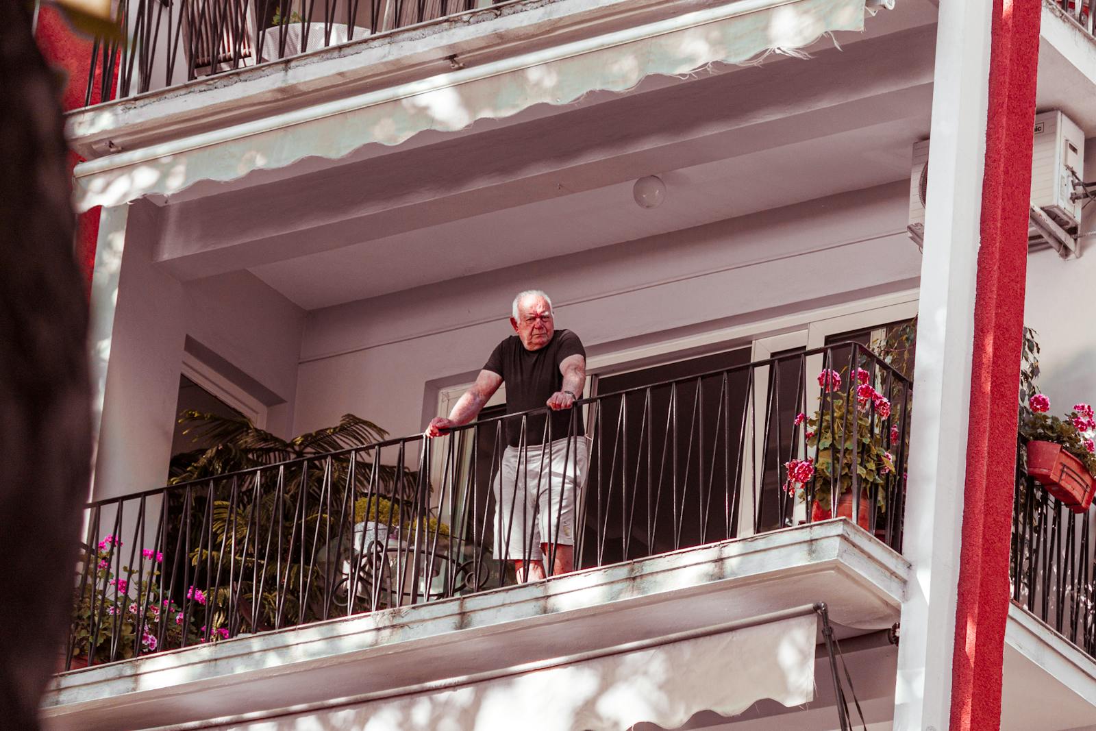 A senior man stands on a balcony surrounded by plants, enjoying a sunny day.