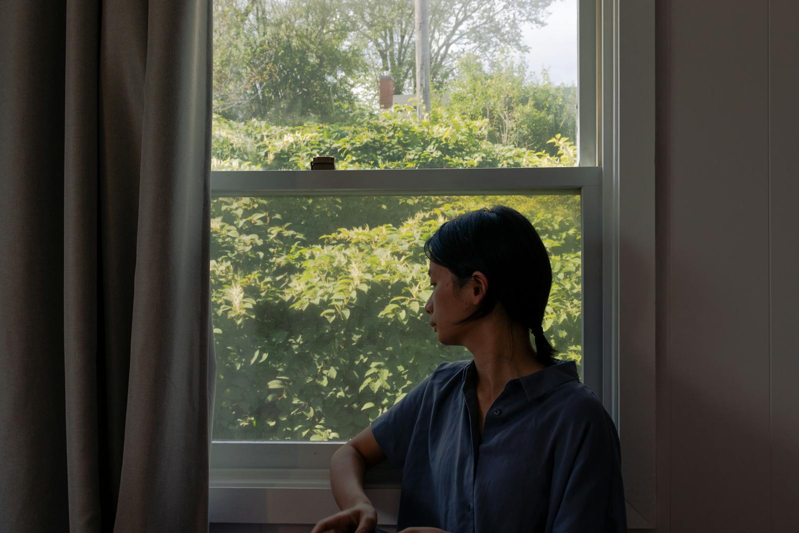 A young woman in casual attire looks outside through a window at lush green foliage.