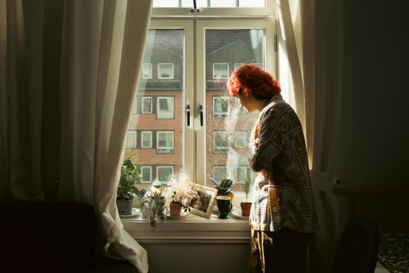 A woman stands thoughtfully by a sunlit window with plants.