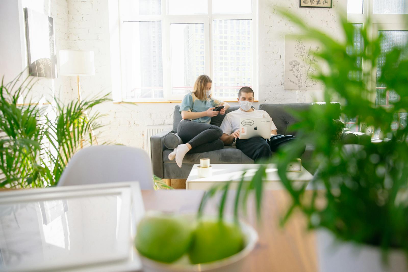 Couple sitting on sofa with masks, using electronics in bright room.