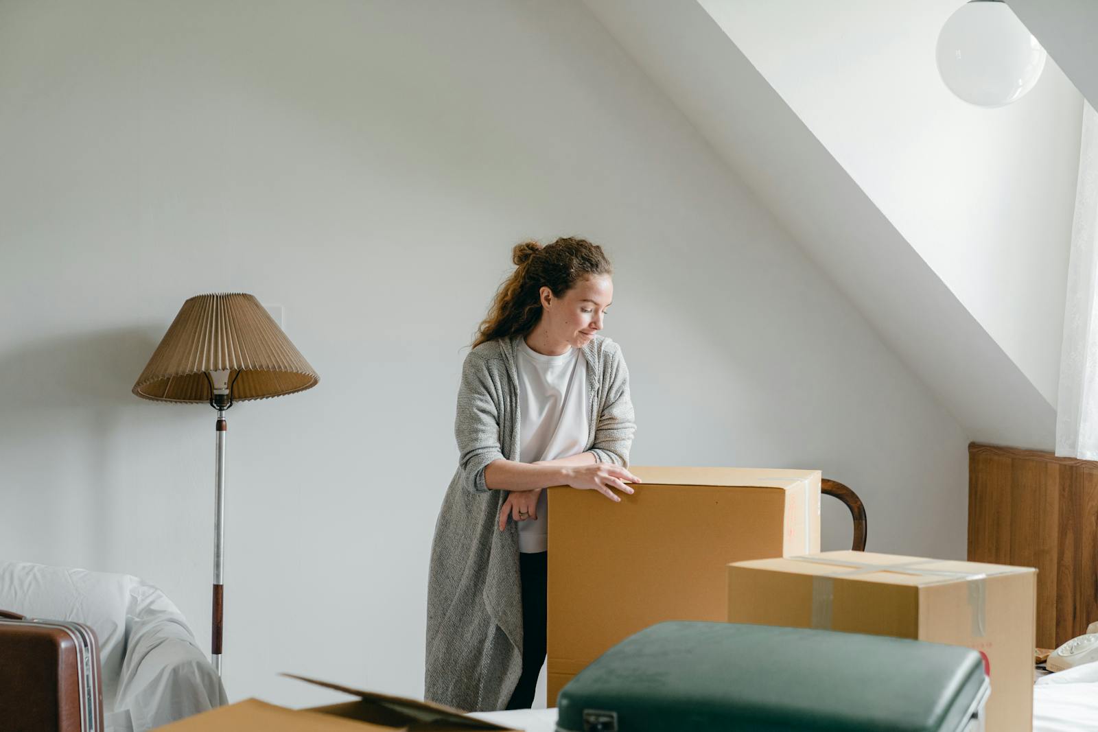 Wistful female in casual wear standing near big cardboard box placed on bed near suitcase on attic style room with floor lamp near sofa and looking down