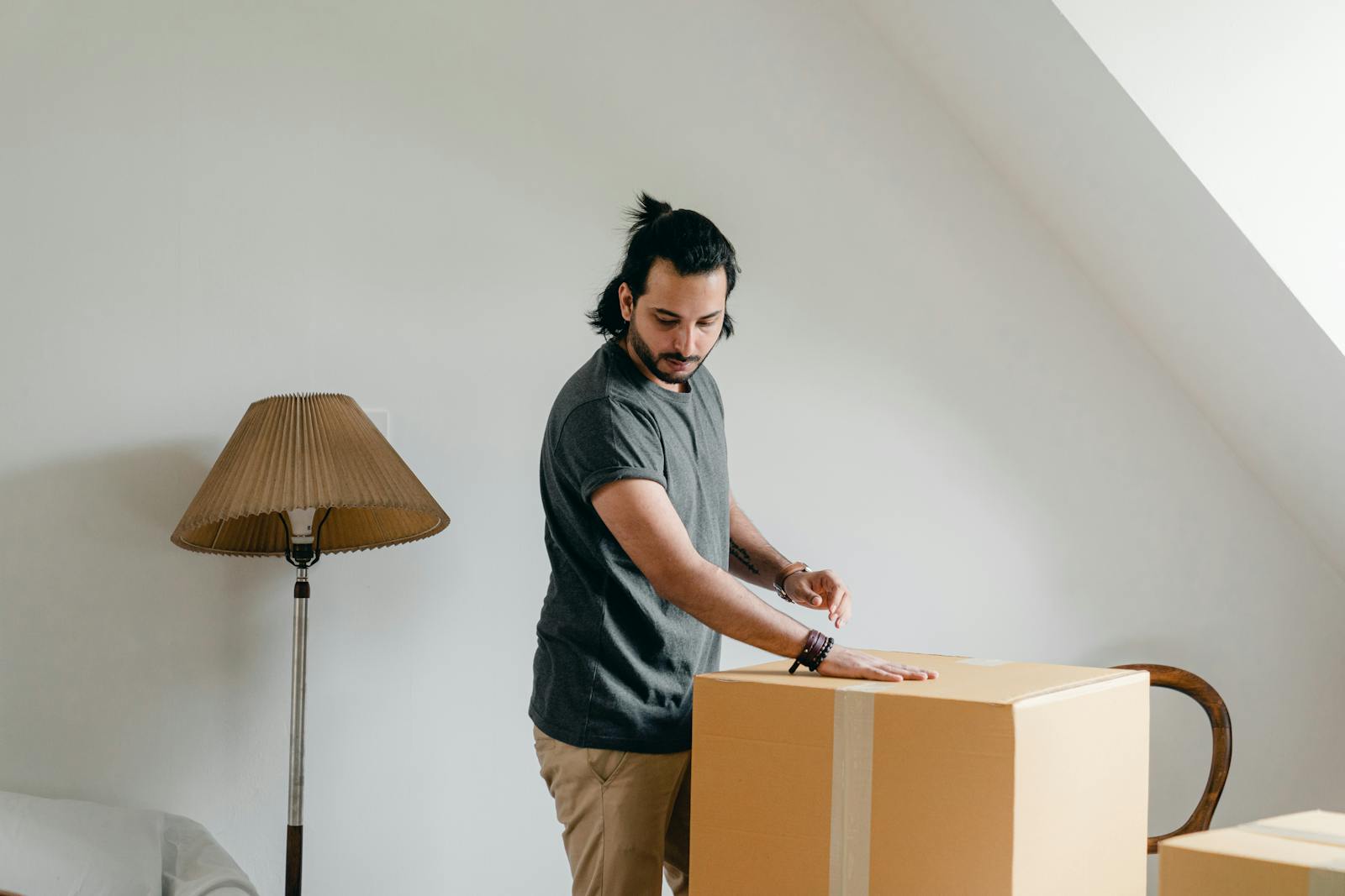 Side view of fit pensive ethnic male in casual wear and wristwatch putting palm of hand onto cardboard box and looking down in attic style apartment