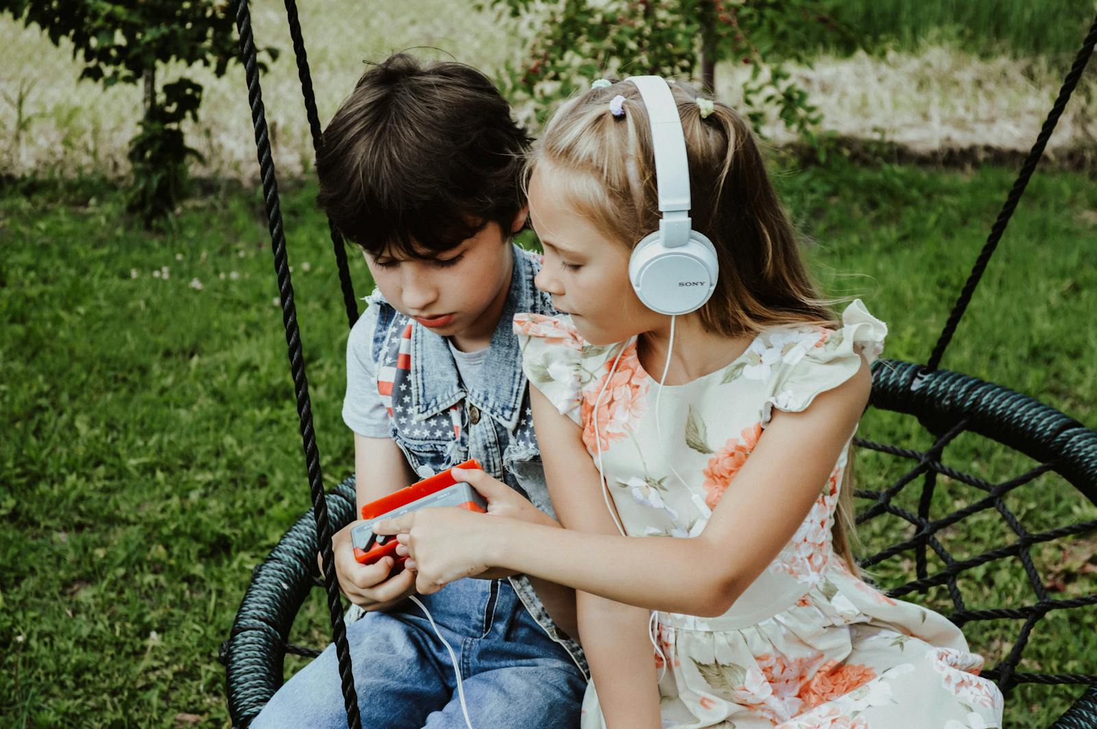 Attentive girl with headphones and serious boy with retro cassette player listening to songs while sitting on swing in grassy park