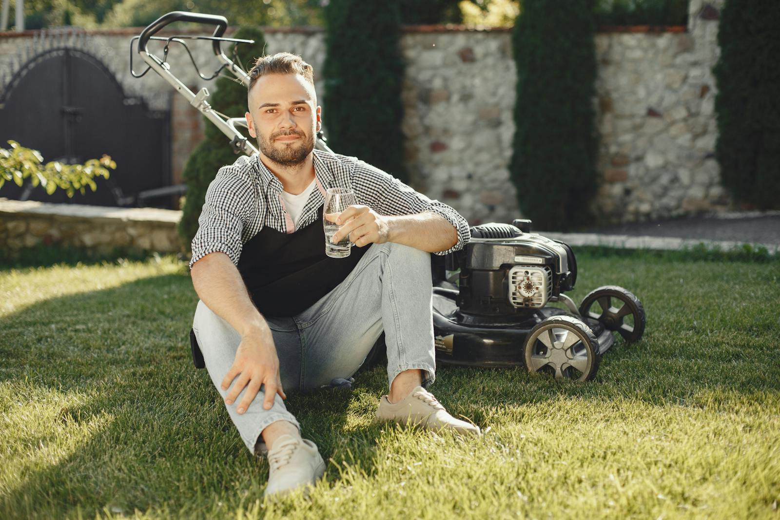 Man sitting on grass with drink, beside lawn mower on a sunny day.