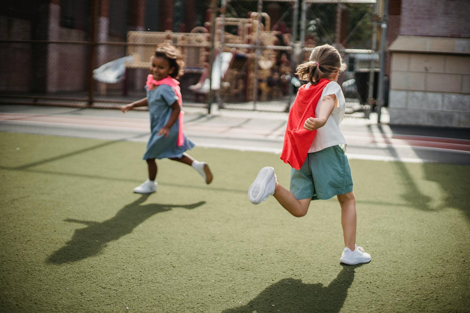 Two children enjoying playtime with capes on an outdoor playground, running on a sunny day.