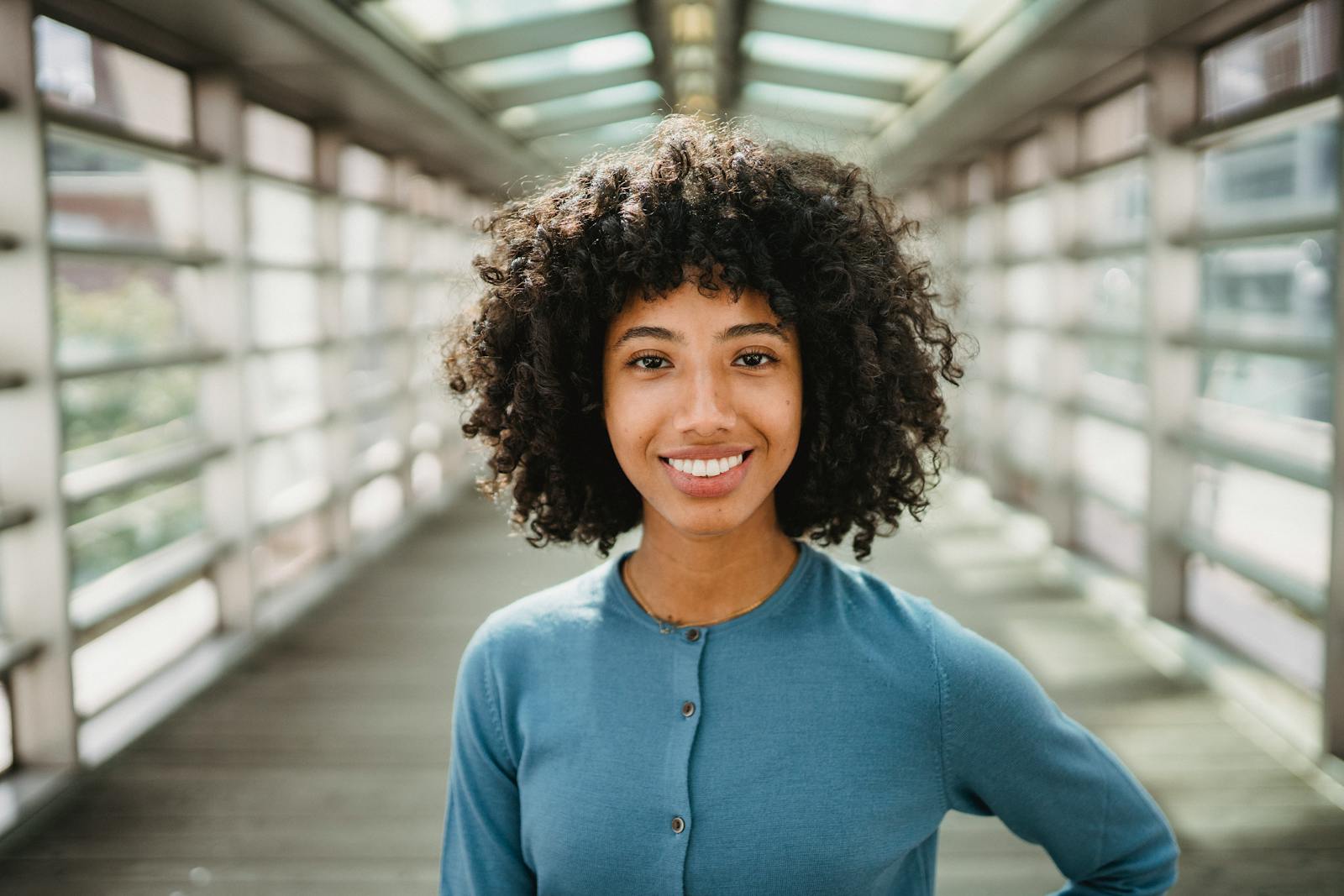 Cheerful African American female with Afro hairstyle smiling and looking at camera while standing on elevated pedestrian crossing with blurred background
