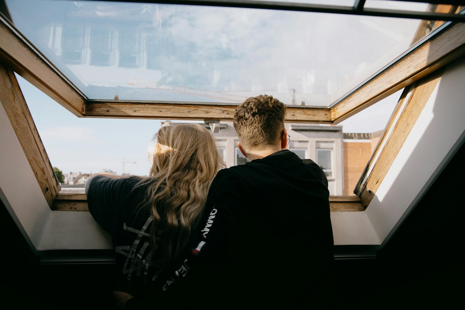 Back view of faceless couple in casual clothes hugging while standing near open window and looking at city