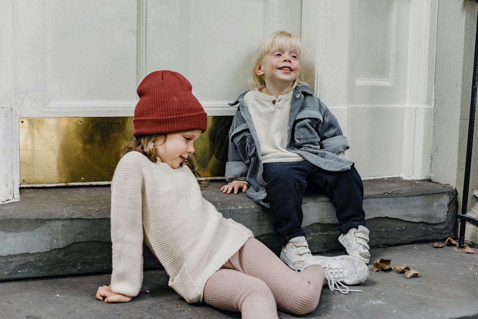 Funny girl in hair and blond boy having fun while sitting on porch near door of residential building on street