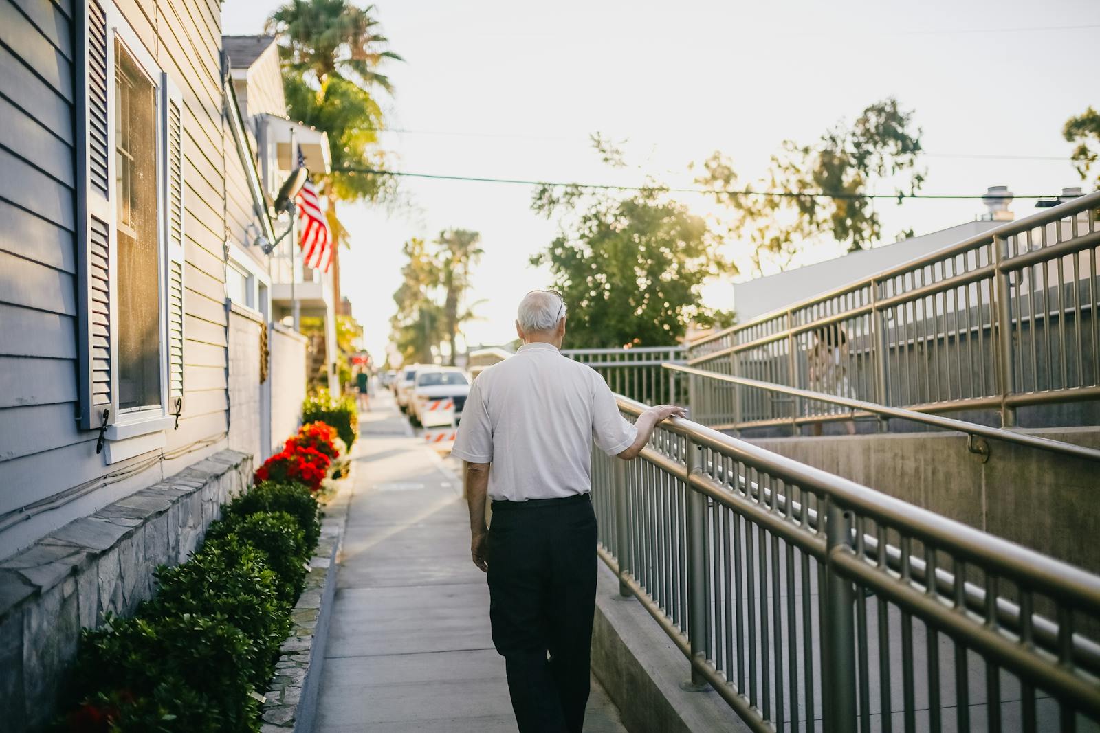 An elderly man walks along a sunny street pathway, surrounded by plants and buildings, capturing a peaceful moment outdoors.