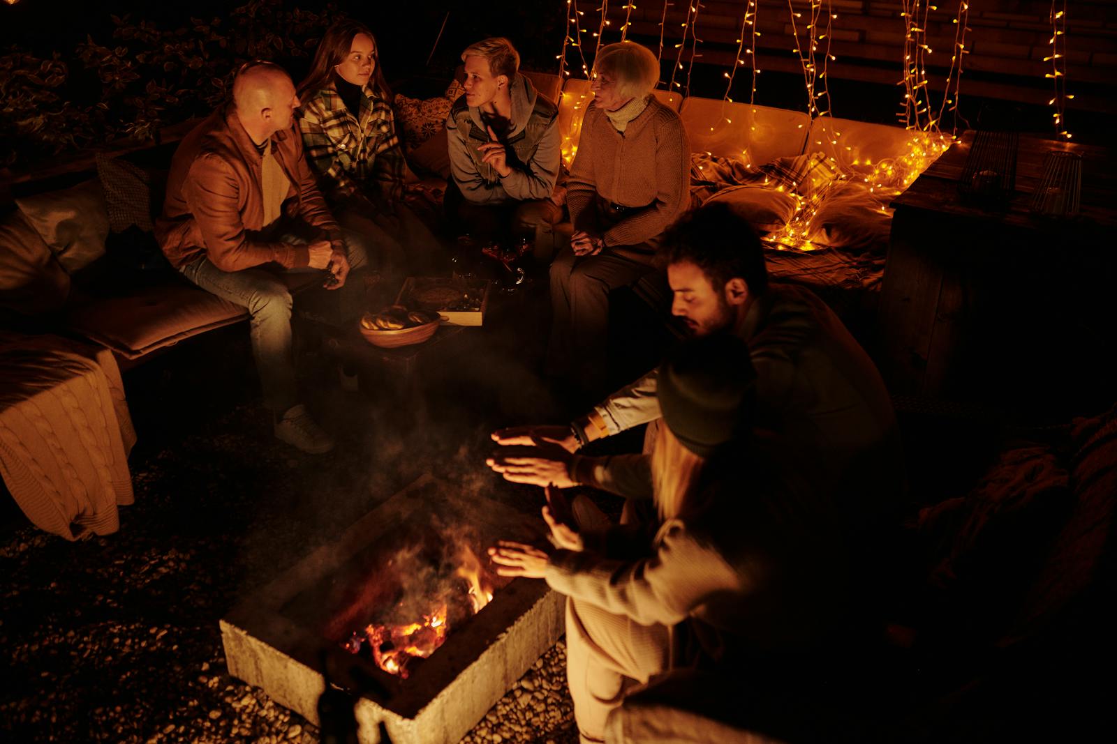 High angle of friends gathering around burning bonfire in garden of house with garlands glowing in dark