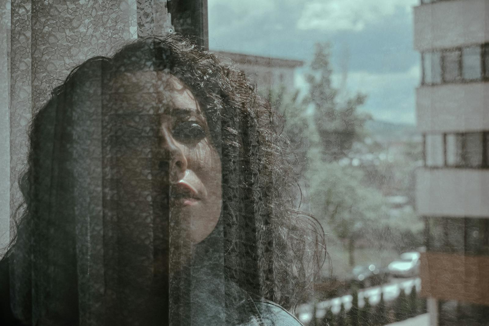Through window of dreamy female with curly hair looking away while standing behind transparent glass with cityscape on background in sunlight