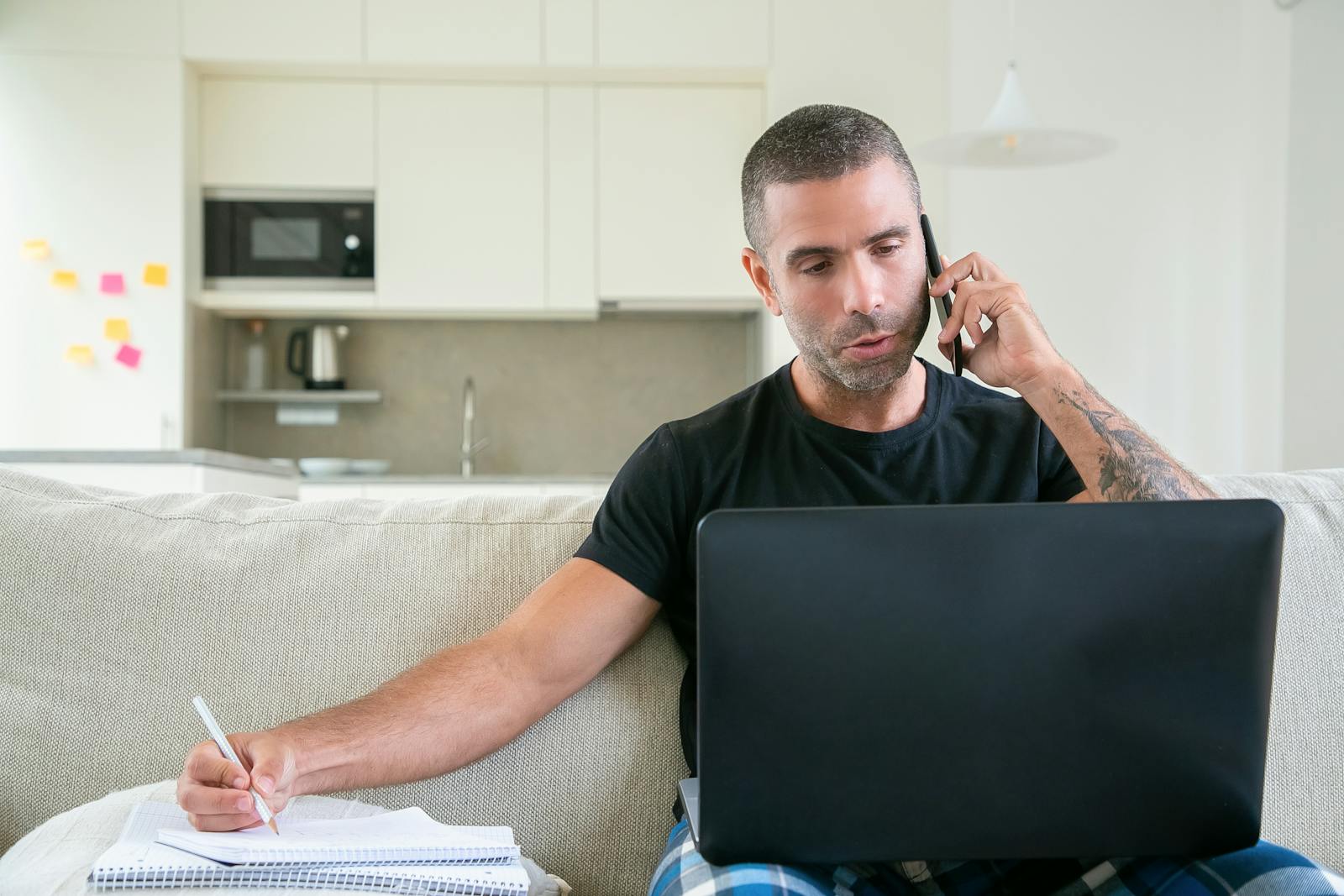 Man working from home using a laptop and smartphone while taking notes in a modern kitchen setting.