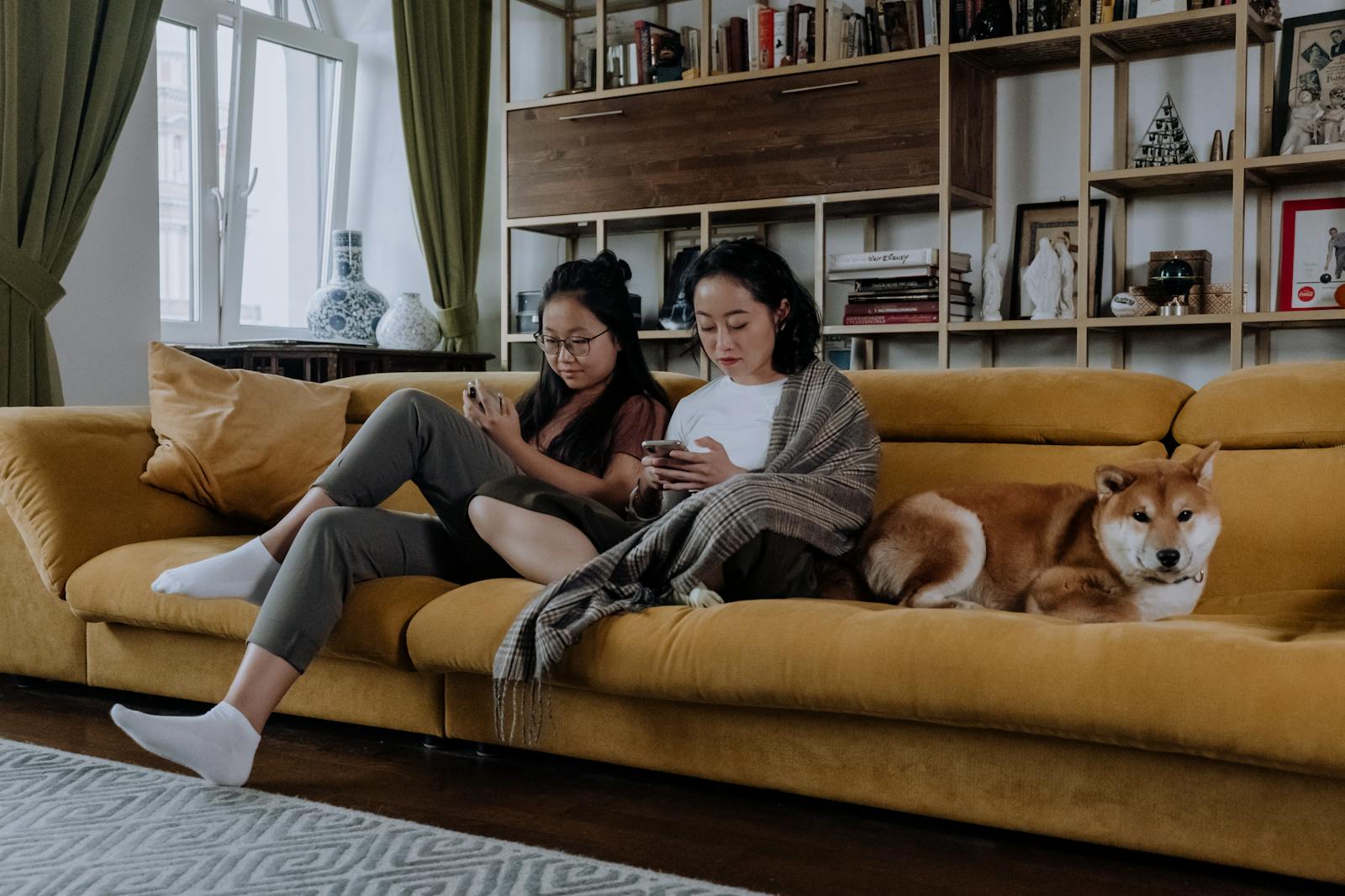 Two women sitting on a sofa with a Shiba Inu dog, using smartphones in a cozy living room.
