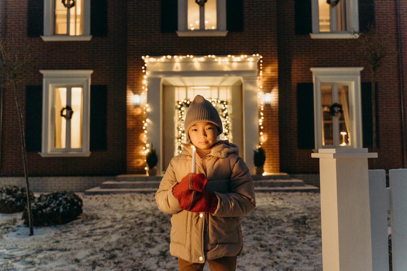 A child in winter clothes holds a candle outside a decorated house at night.
