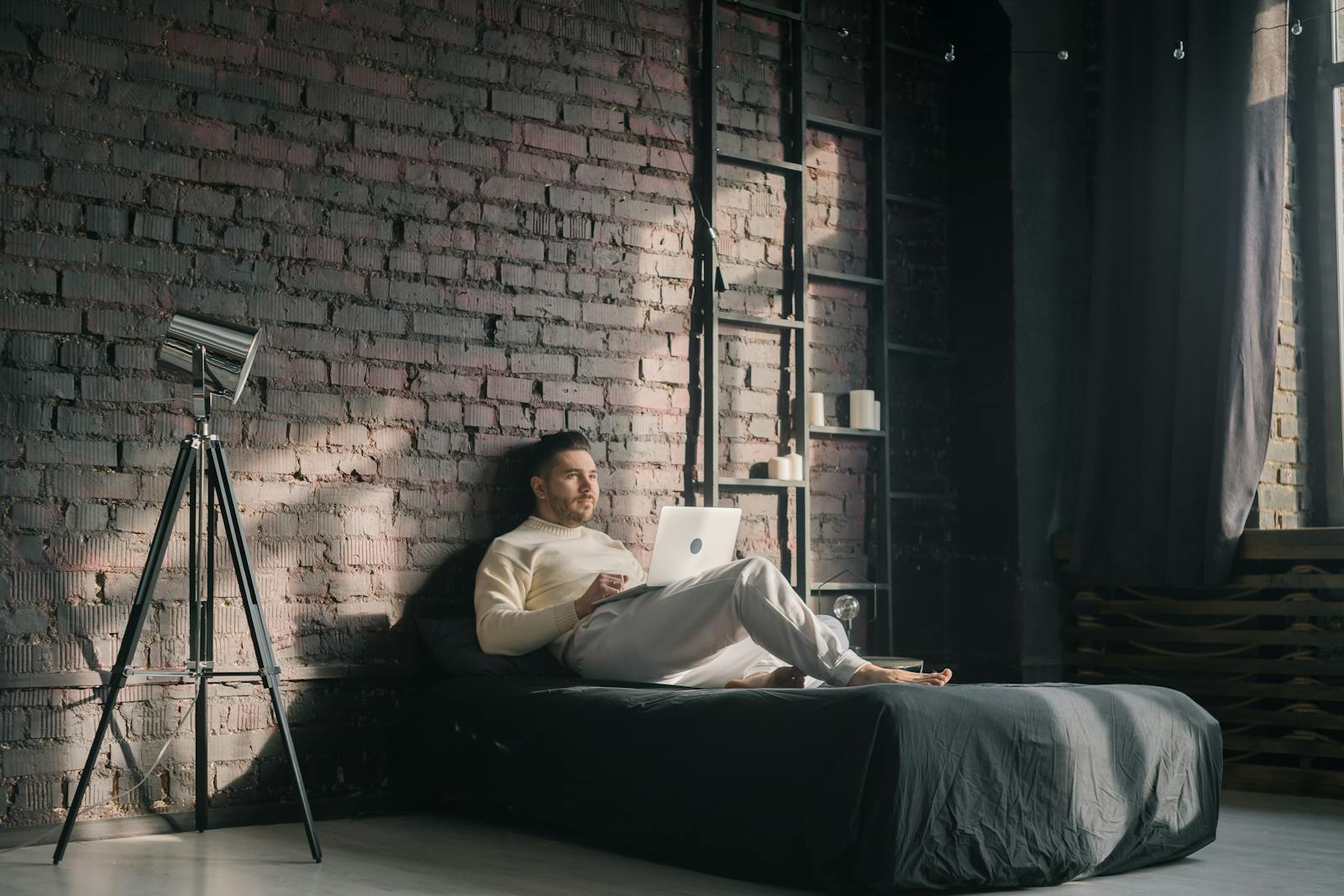 A man lounging on a bed using a laptop in a modern loft setting with natural light.