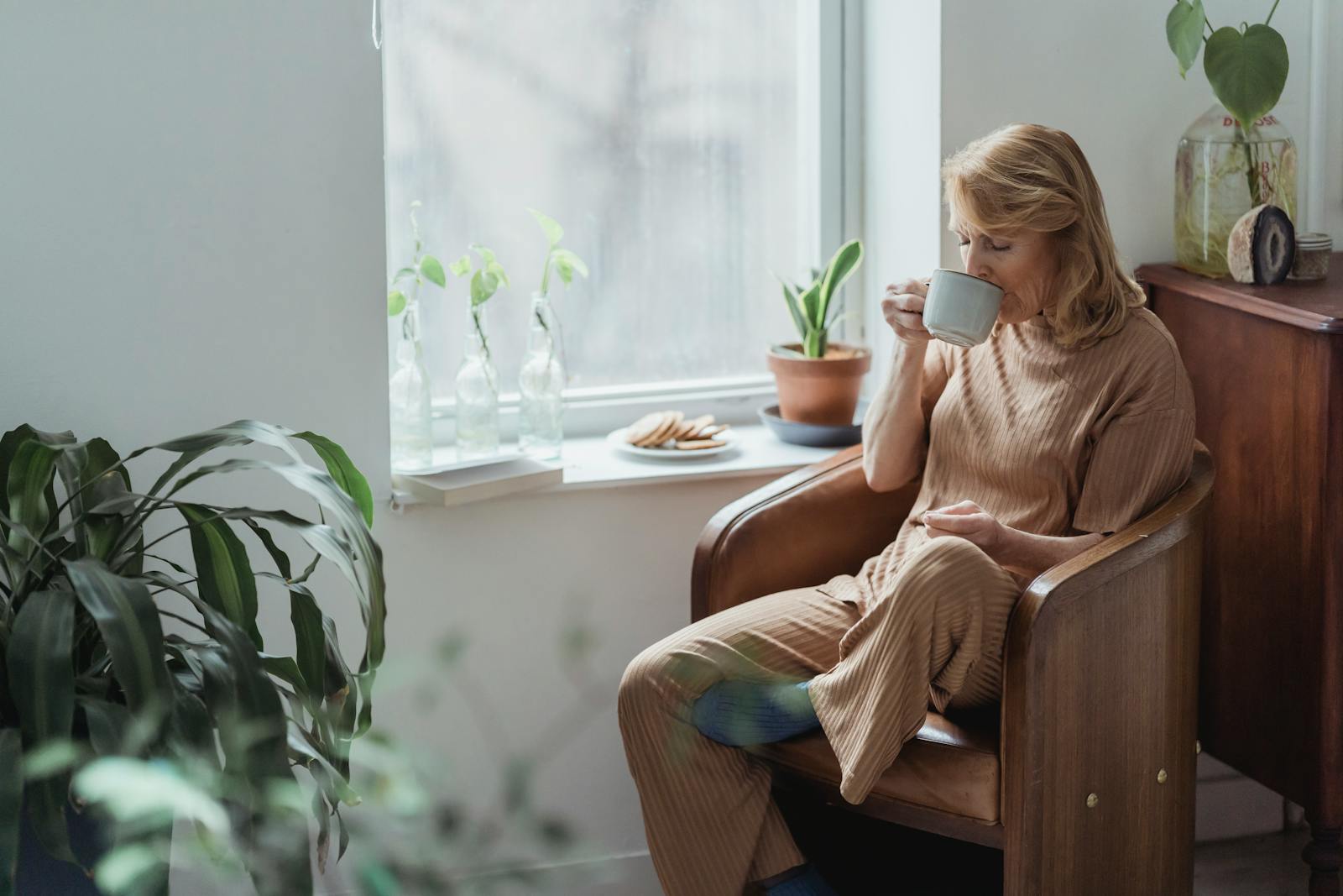 A woman enjoys a peaceful moment with coffee in a cozy, plant-filled living room by the window.