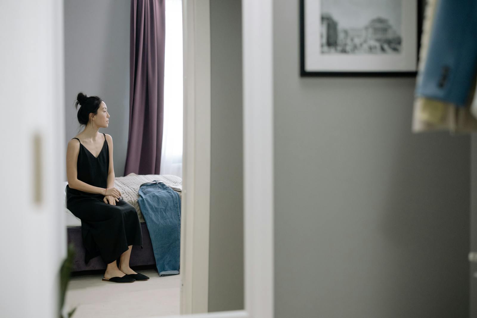 Woman in black dress sitting on bed in a minimalist bedroom setting, reflecting a calm and serene atmosphere.