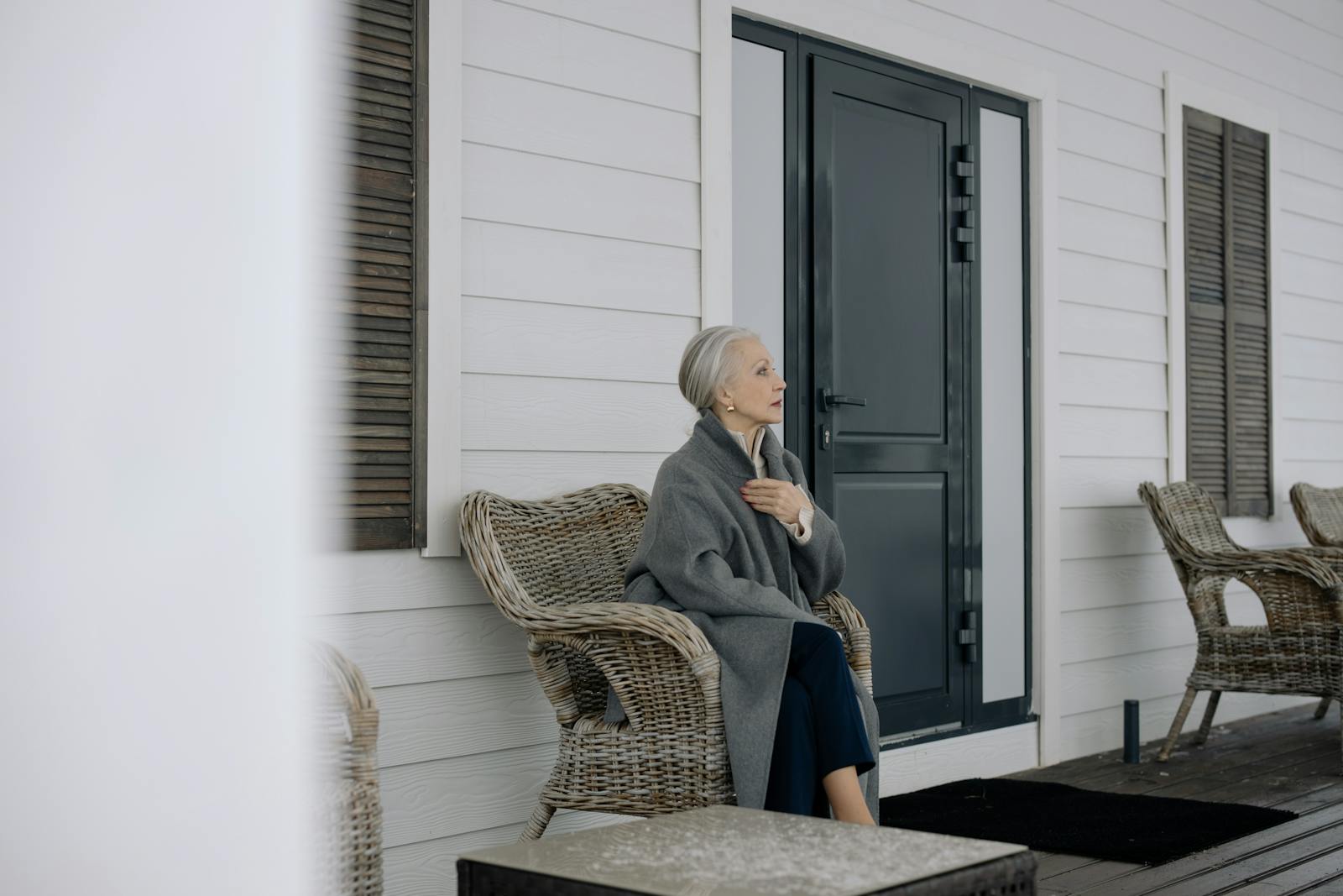 Elderly woman sitting on a wicker chair on a porch, wrapped in a blanket, during wintertime.