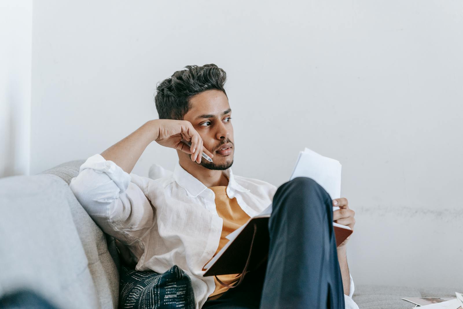 Contemplative young ethnic male in casual wear reading notes in planner and looking away in thoughts while resting on comfortable couch