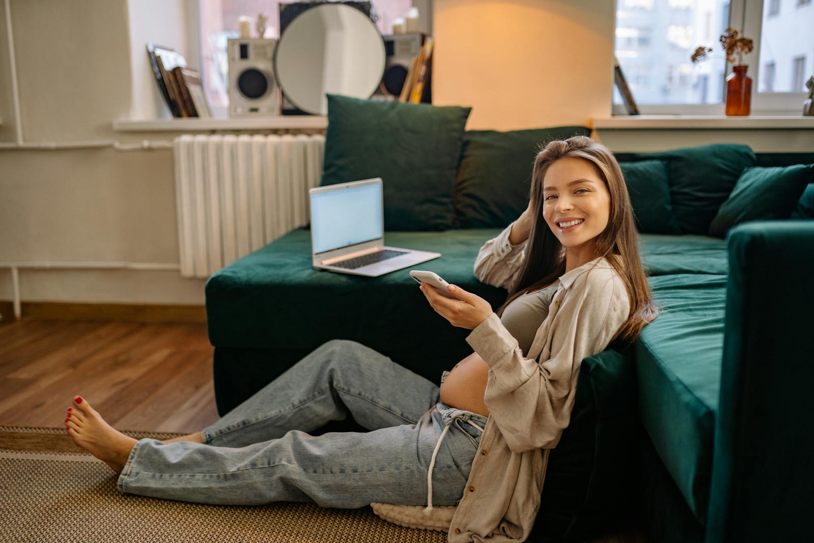 Smiling pregnant woman sitting on the floor using smartphone indoors.