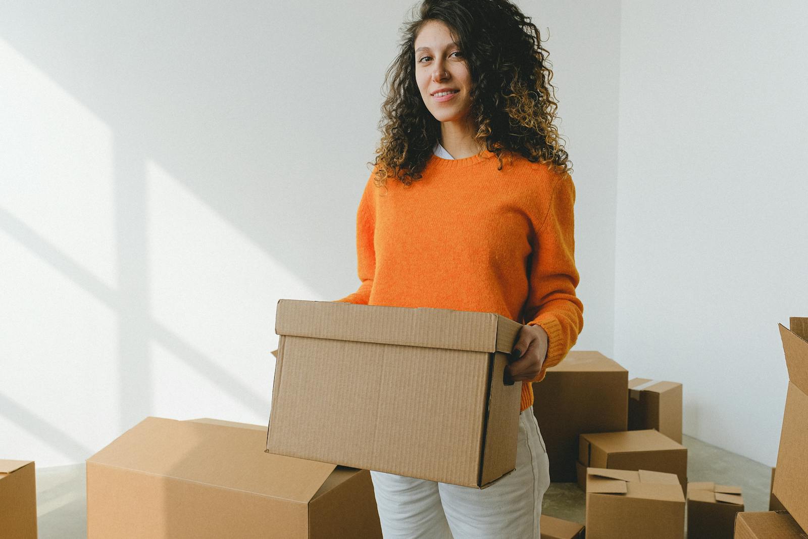 Positive young lady with dark curly hair in stylish orange sweater holding carton box and looking at camera during relocation in daylight