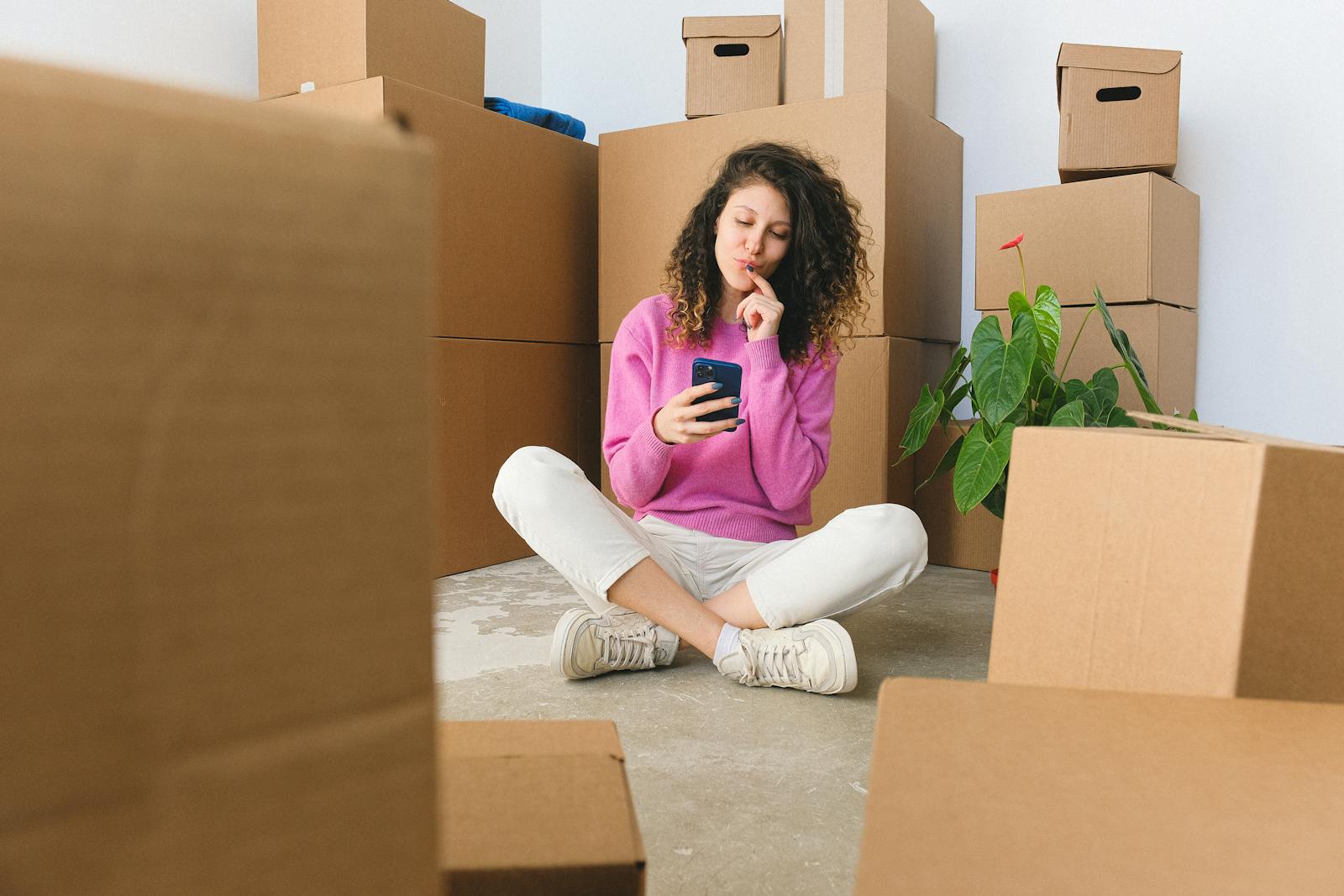 Full body of positive thoughtful young female millennial in casual outfit touching lips and using smartphone while sitting on floor with crossed legs near carton boxes after relocation