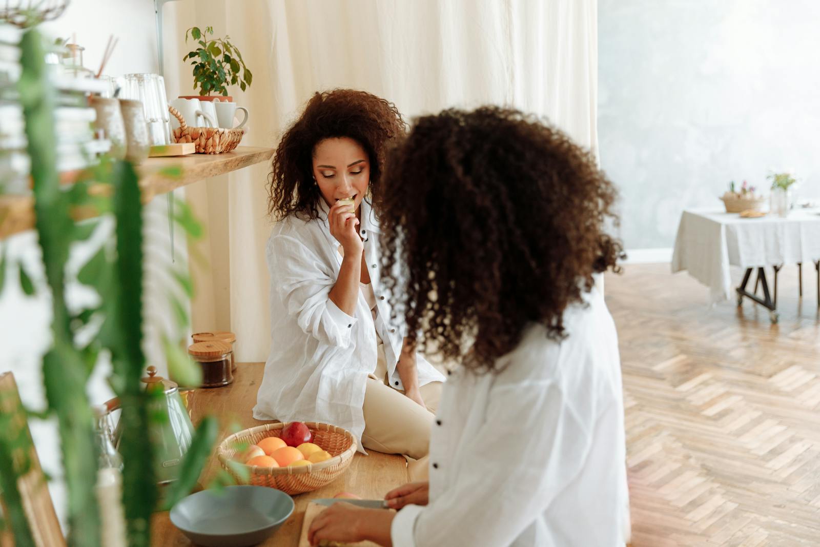 Two women enjoying a casual, stylish moment in a cozy kitchen setting with fresh fruits.