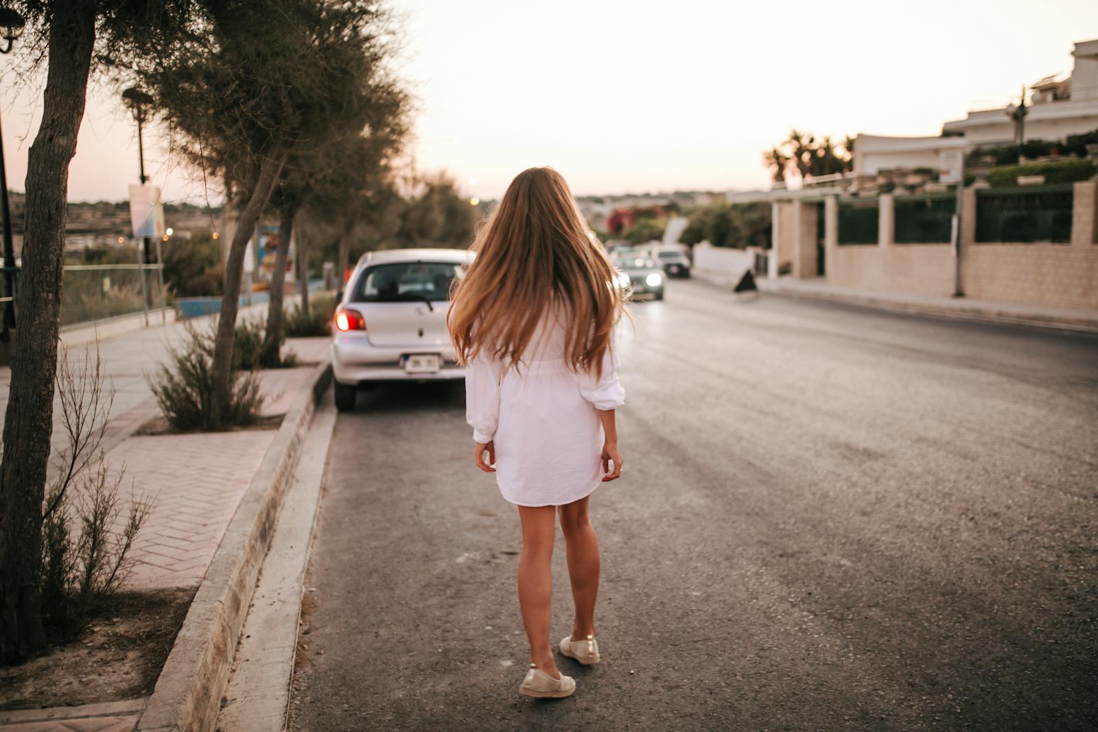 A woman in a white dress walks along a quiet suburban street at sunset, surrounded by parked cars and trees.