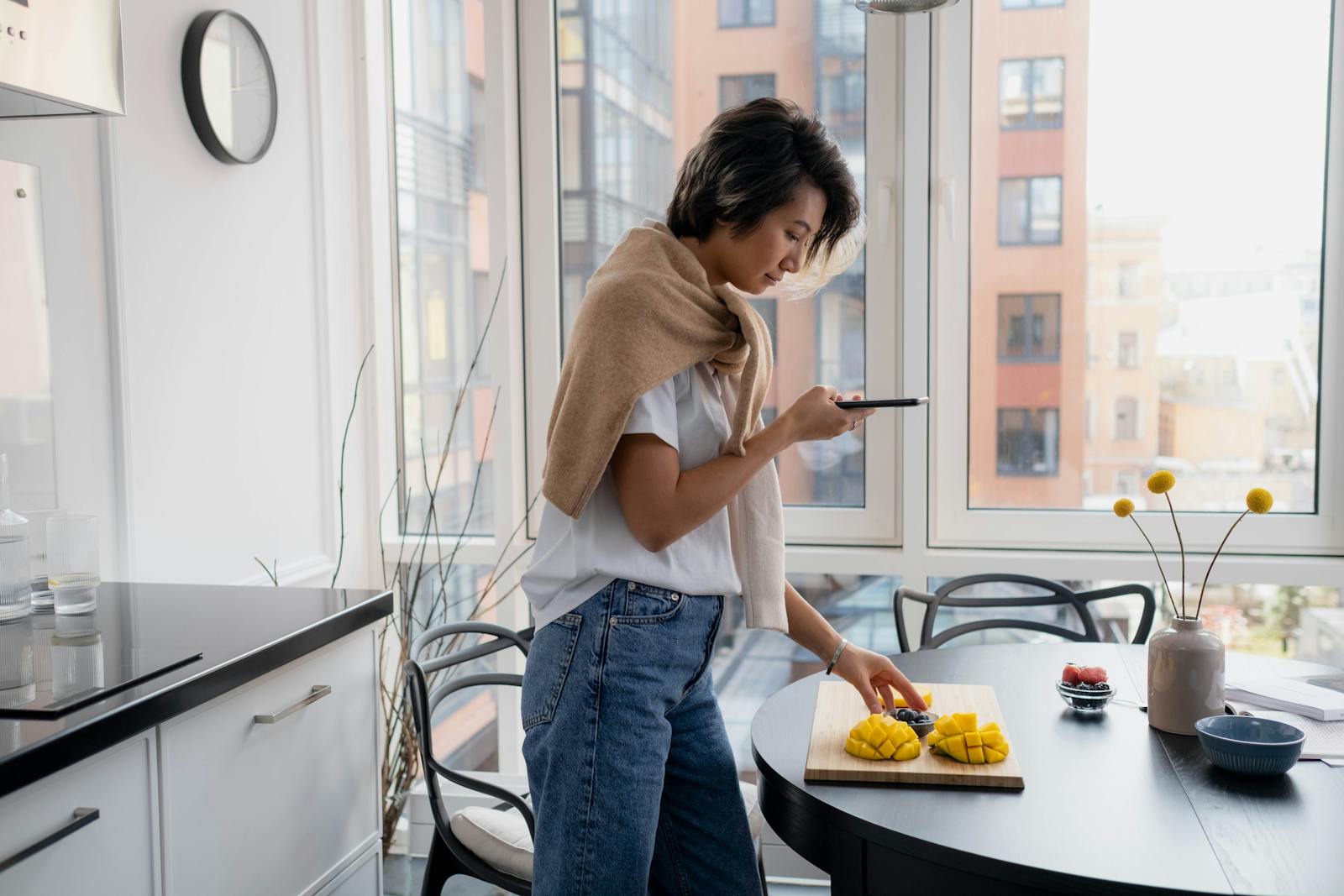 Young woman uses smartphone to photograph a cutting board with fresh mango in modern kitchen.