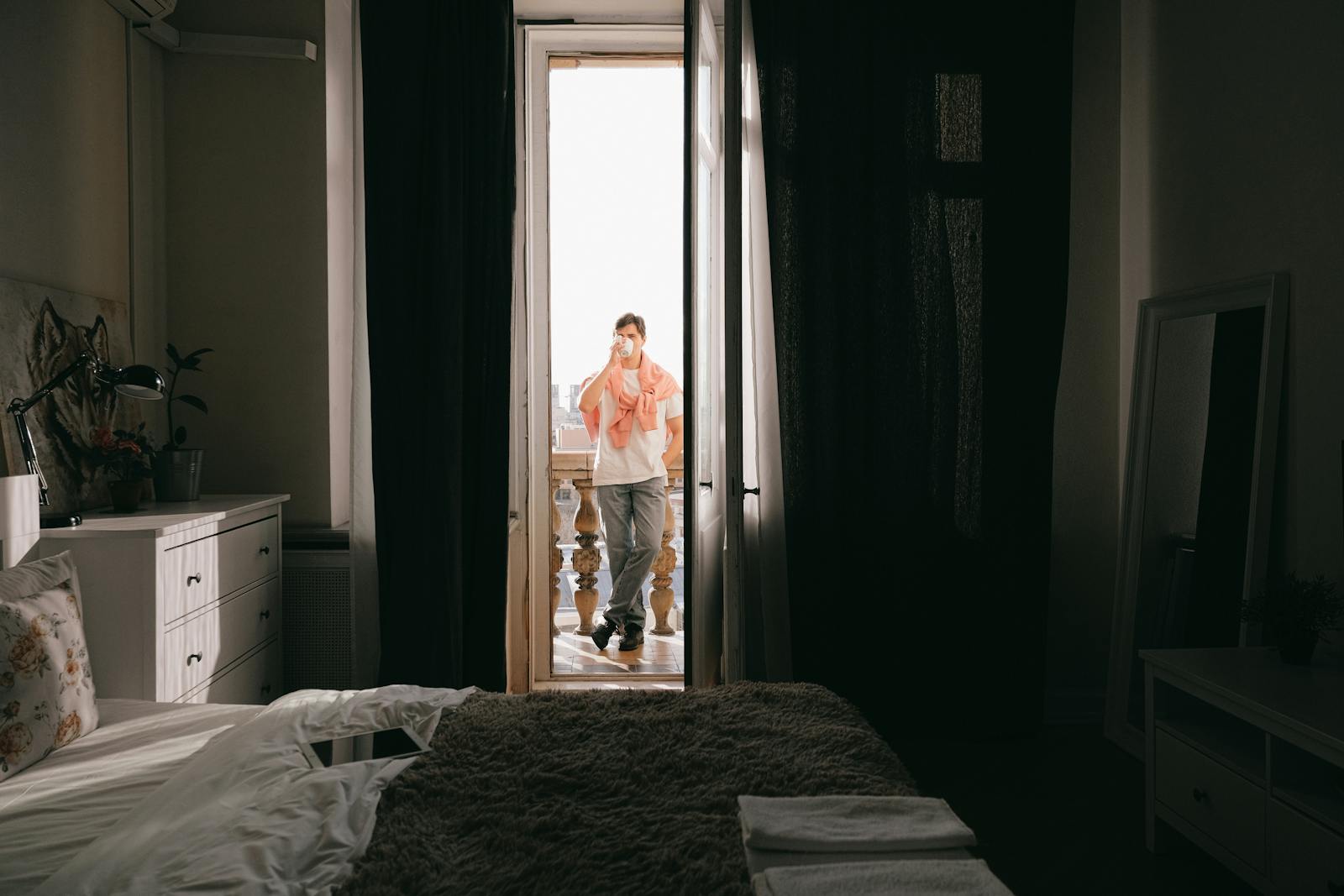 A man stands on a balcony enjoying coffee, morning light streaming into a cozy bedroom.