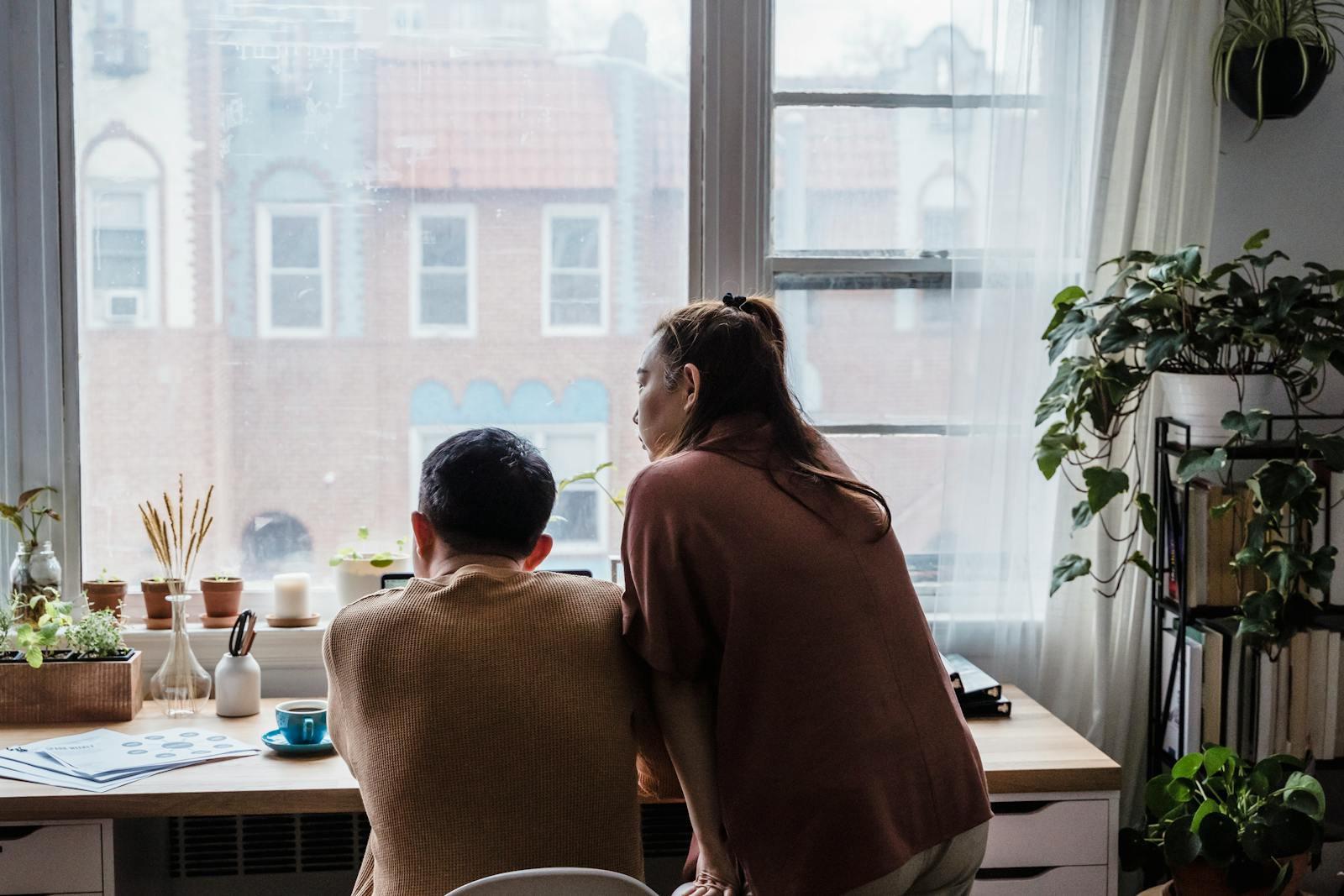 A couple enjoys the view outside their home office window, surrounded by plants.