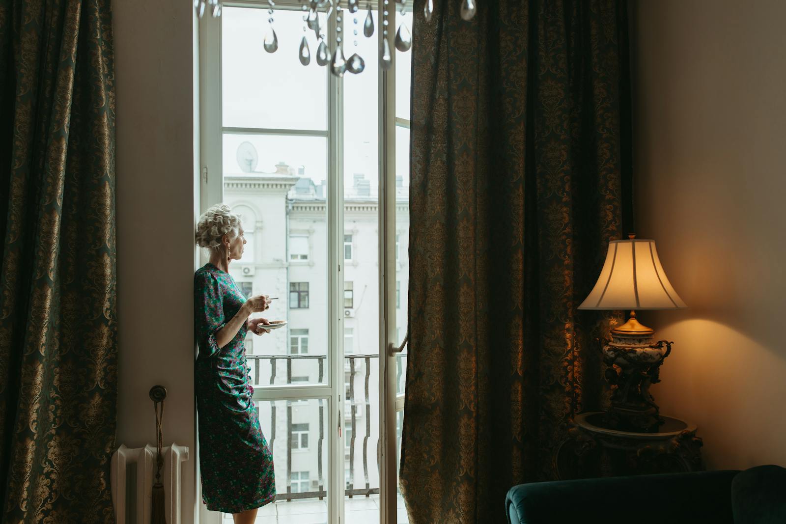 A woman stands by a glass door in an elegantly decorated room with curtains and a lamp.
