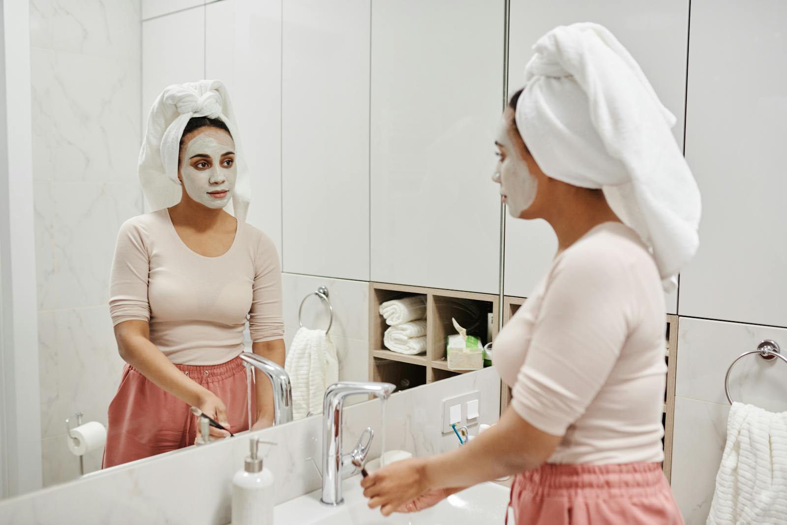 Adult woman with clay mask and towel relaxed in modern bathroom mirror reflection.