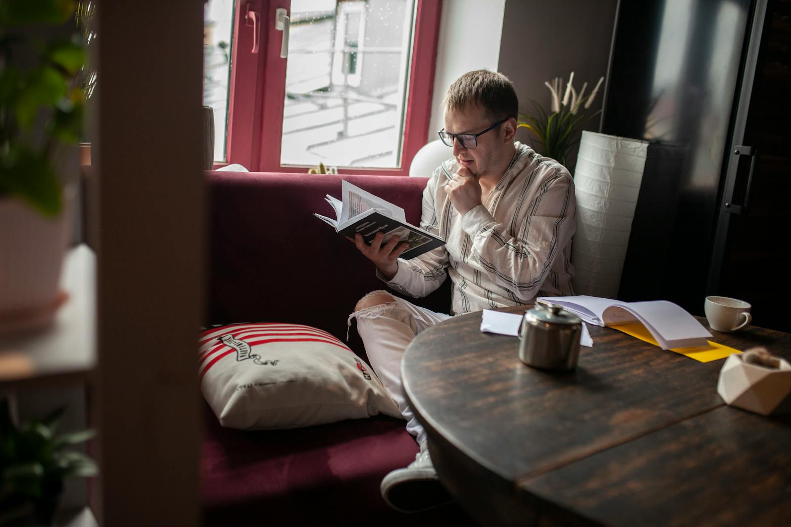 A man sits in a cozy room, reading a book by the window with a thoughtful expression.