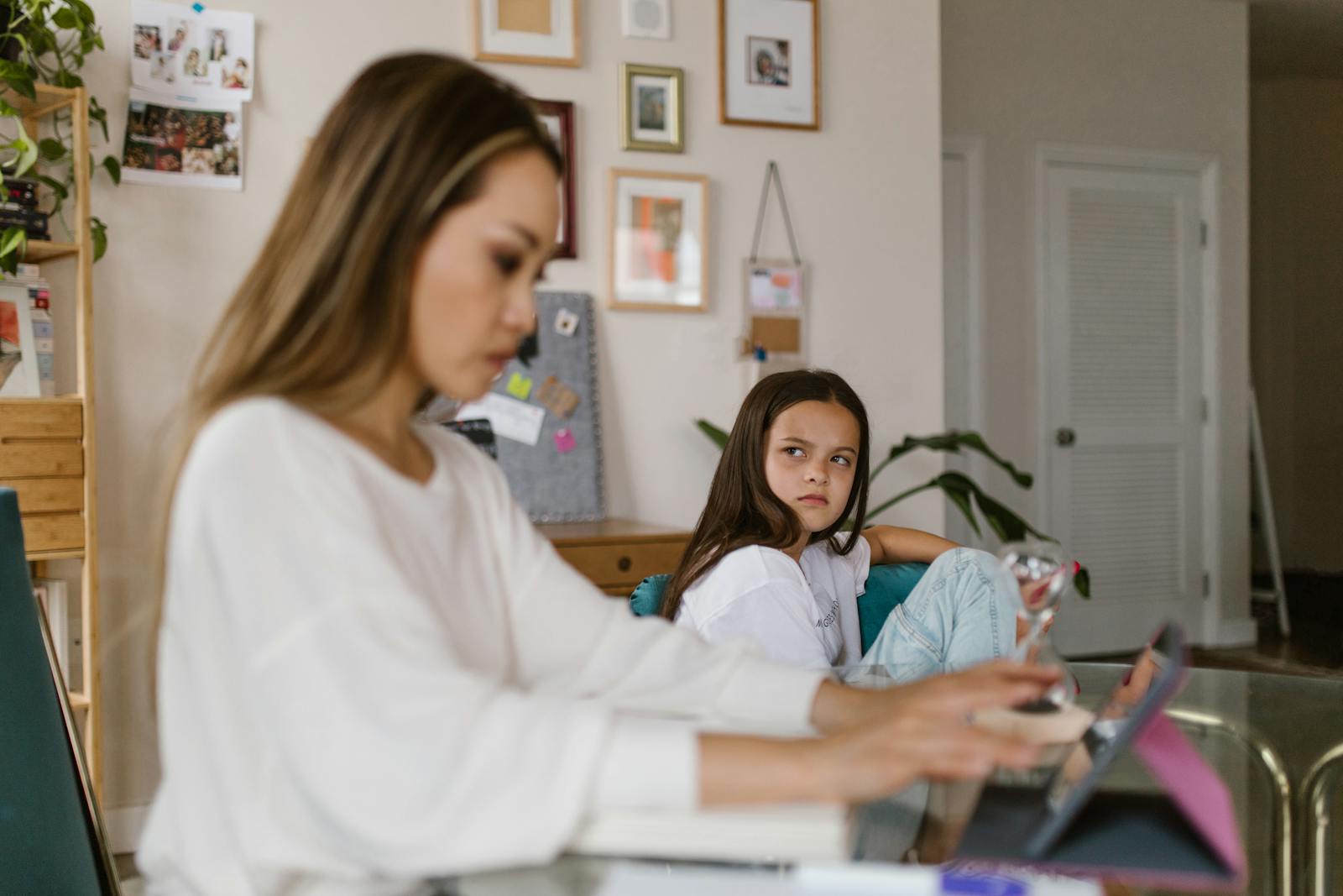 A woman working on a tablet at home while a young girl looks on, creating a work-from-home family setting.
