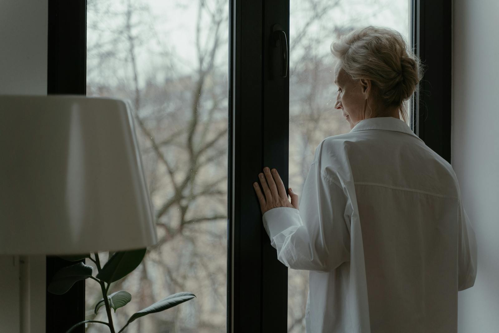 A senior woman in white looking out a window thoughtfully, symbolizing reflection.