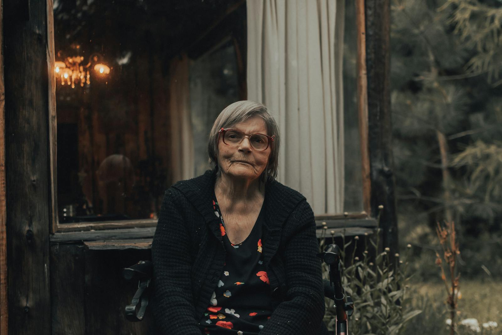 Senior woman in a cardigan with eyeglasses sitting outside a rustic cabin.