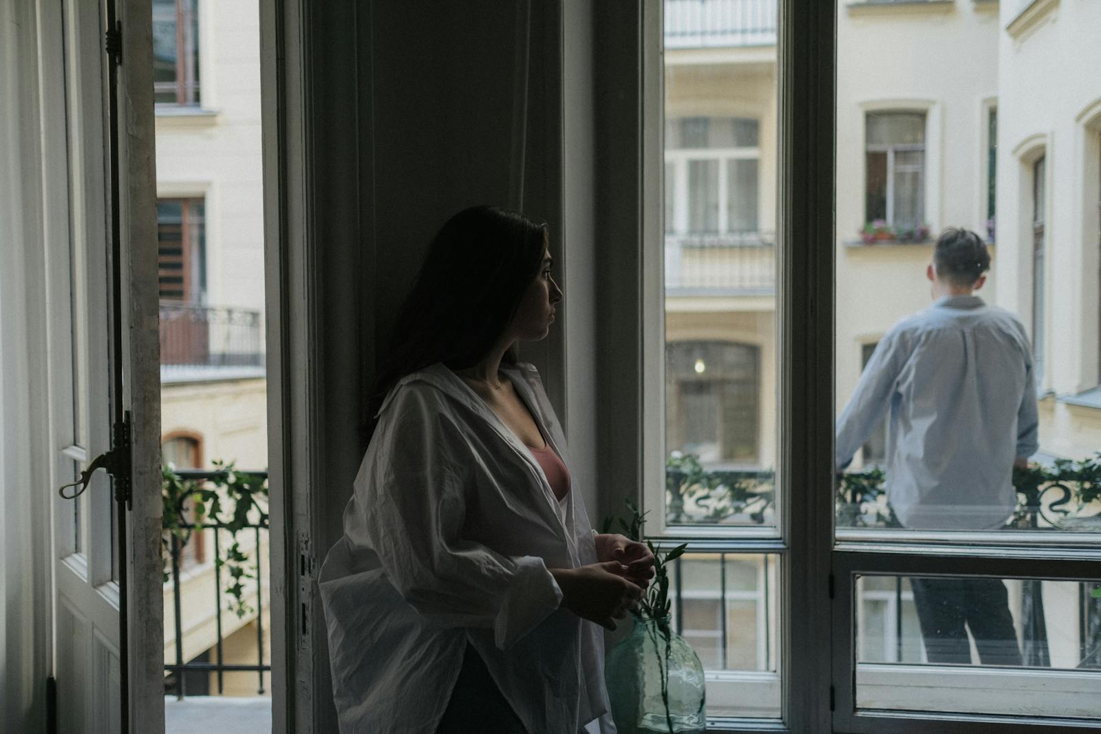 A couple stands indoors and on a balcony, creating a scene of intimacy and urban life.