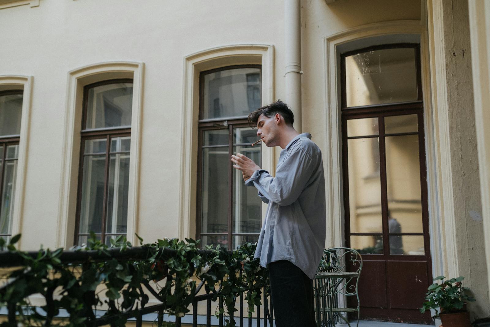 A man stands on a balcony, smoking and looking contemplative in an urban setting.