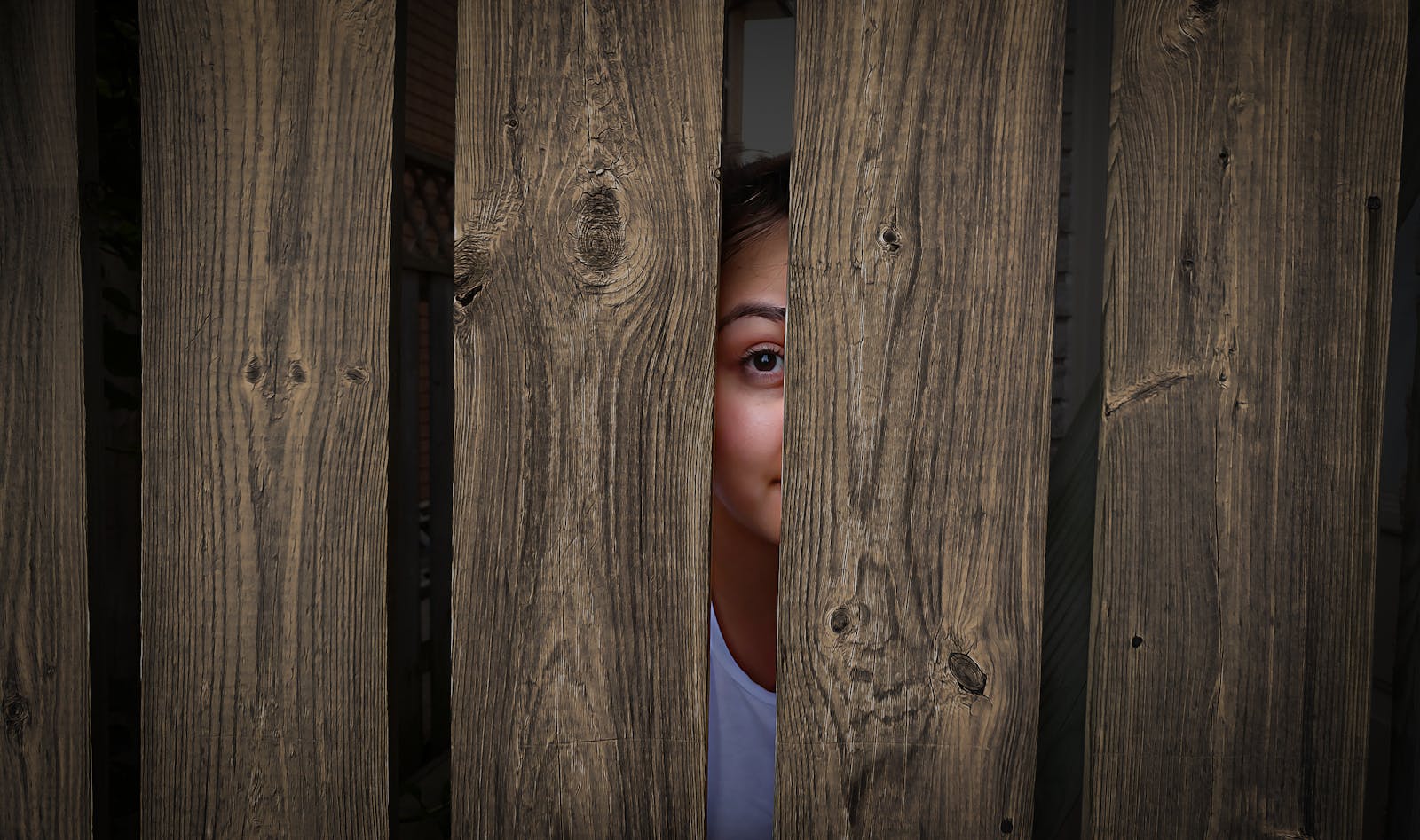 A girl peeks curiously through a rustic wooden fence, capturing a moment of curiosity and mystery.