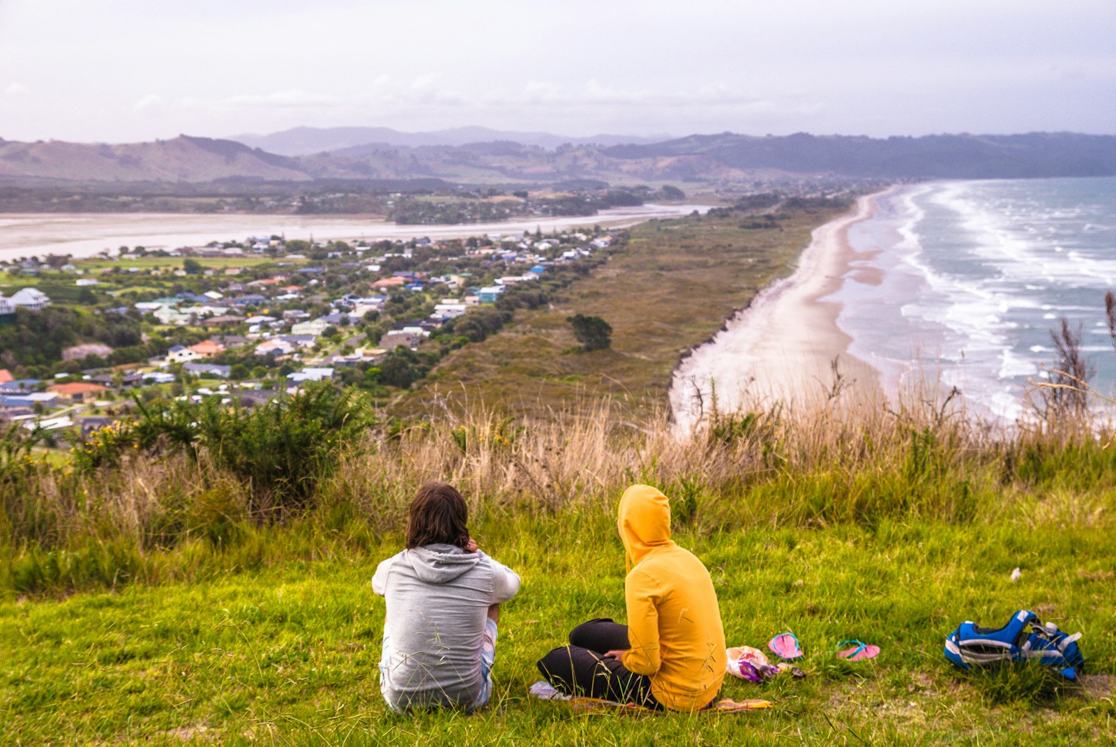 two people sitting on a hill overlooking a beach and town