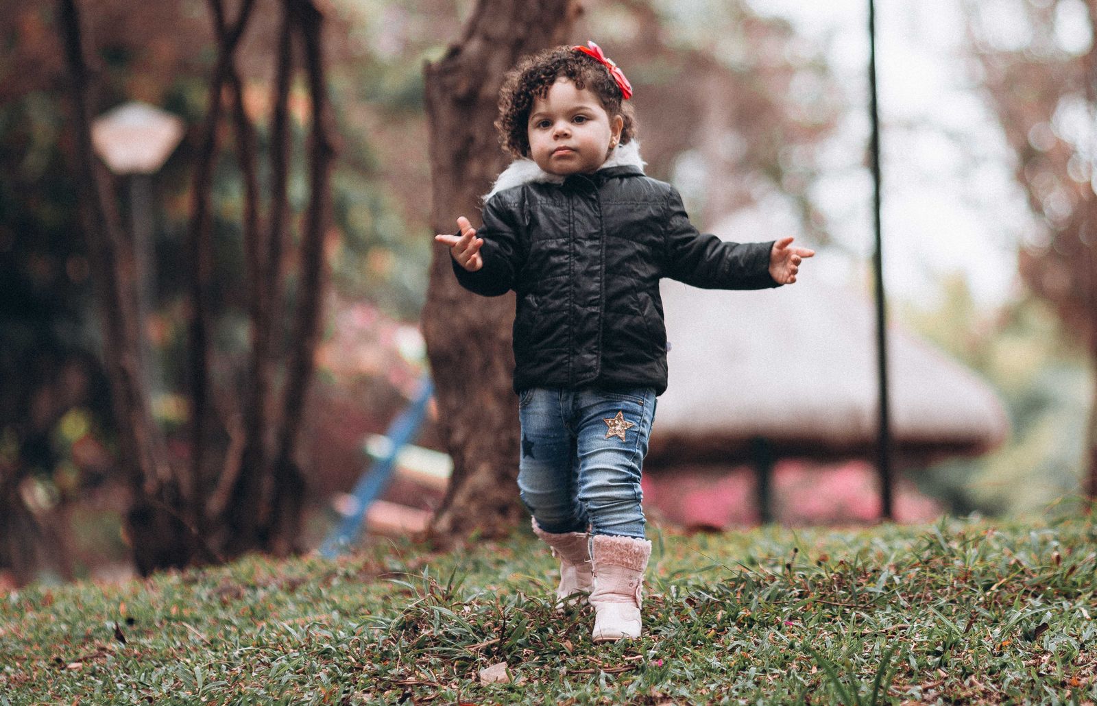 A young girl wearing a jacket walks on grass in an autumn park setting.
