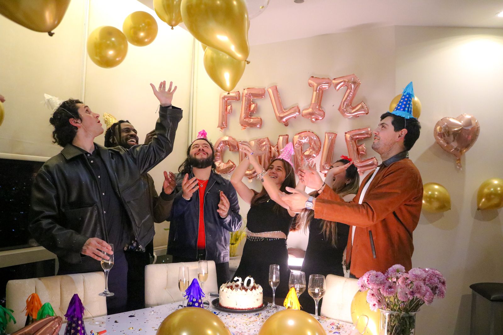 Group of friends celebrating a birthday indoors with balloons and cake.