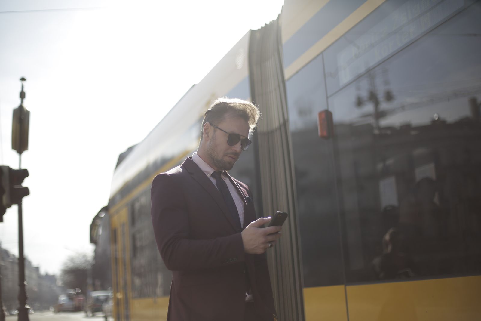 Business professional checks phone at a sunny train station, wearing a suit and sunglasses.