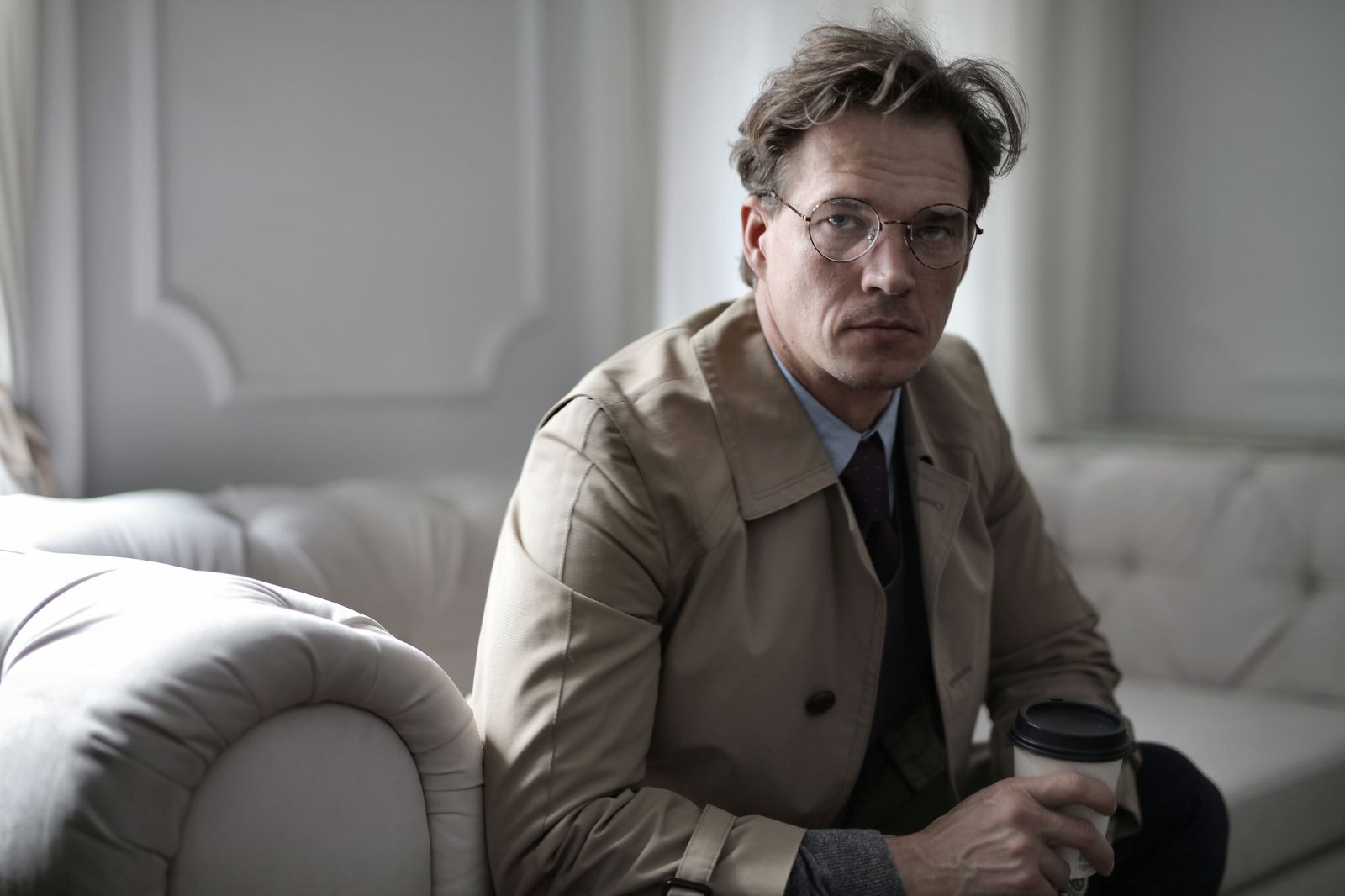 Tired adult male in trench with tousled hair sitting on sofa with paper cup of coffee and looking at camera