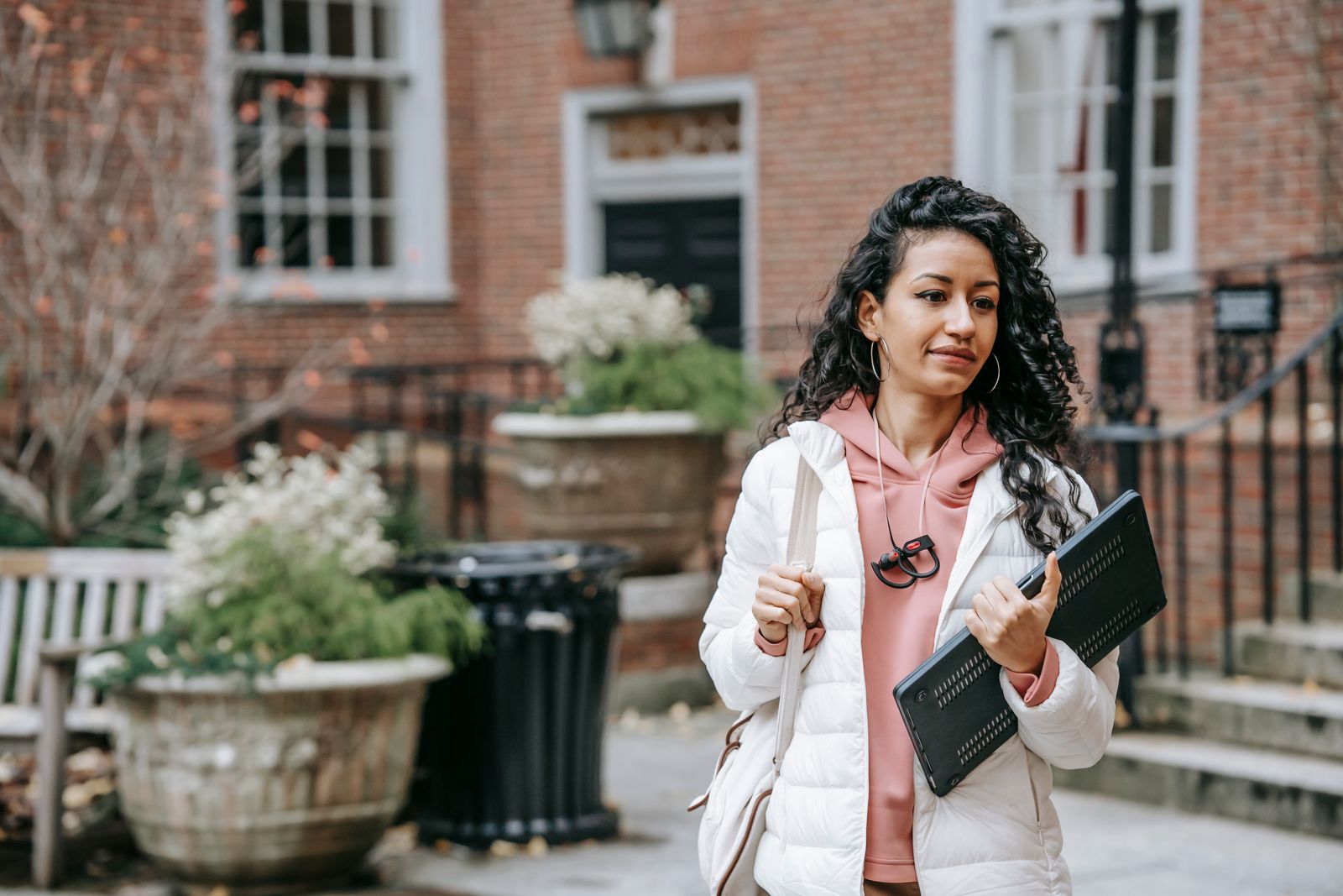 Young ethnic female with long curly hair and netbook in casual trendy outfit on blurred background of brick building