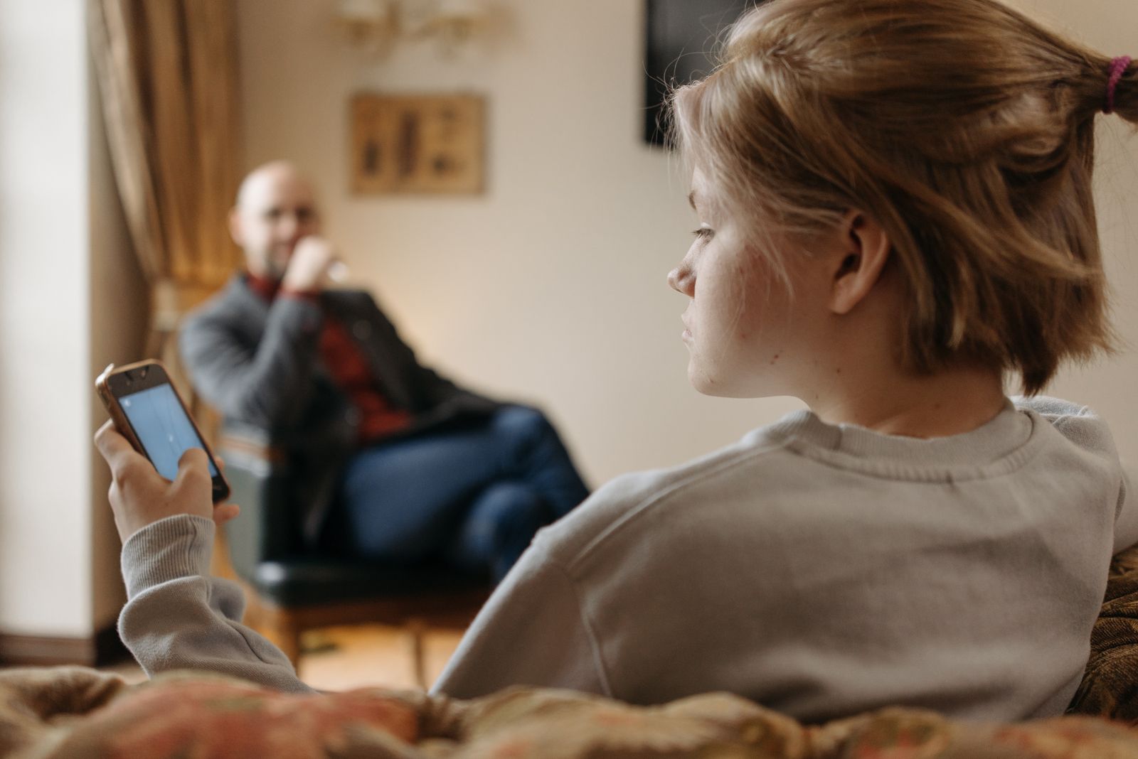A teenager looks at a smartphone while sitting indoors with an adult in the background.
